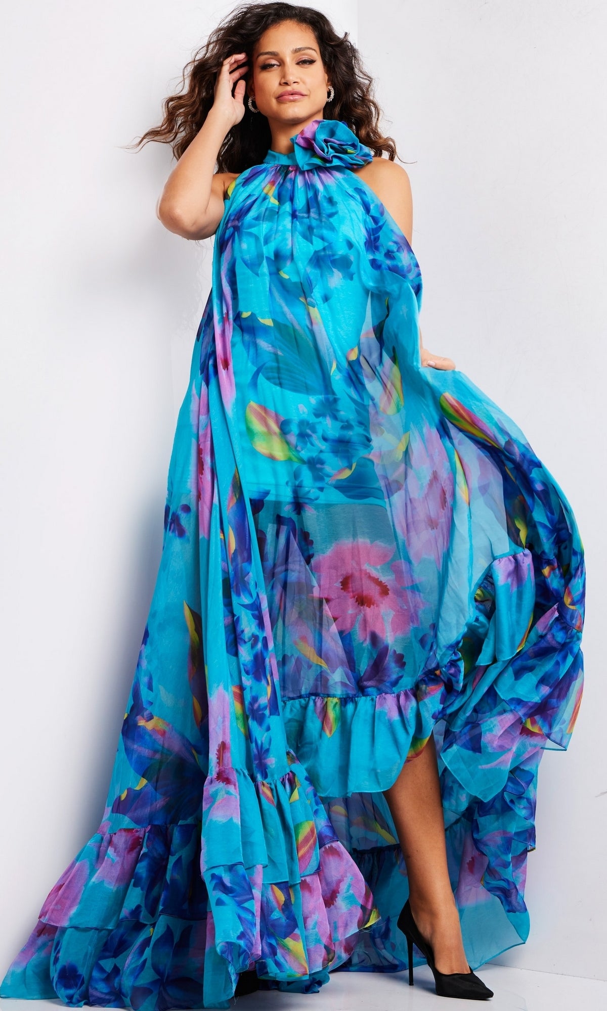 A woman poses in the Long Prom Dress 38721 by Jovani, a bright blue chiffon formal gown with a high neckline, ruffled hem, and sheer fabric, paired with black heels against a plain white background.